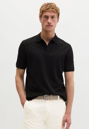 Blondyn ubrany w czarną koszulkę polo z krótkim rękawem, białe spodnie i pleciony beżowy pasek, stoi z jedną ręką w kieszeni.