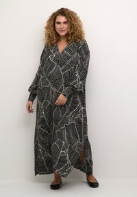 Maxi dress in black with a white leaf pattern. Long sleeves, V-neckline, and side slits. Soft fabric with a flowing silhouette. Black flats.