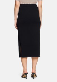 Femme portant une jupe crayon midi noire taille haute avec une fente sur le côté gauche, associée à des sandales à talons noires.