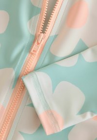 Combinaison de surf pour enfants en bleu clair avec de grands motifs floraux pastel roses et blancs. Dotée d'une fermeture éclair couleur pêche et d'une texture de tissu lisse.