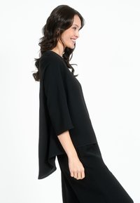 Maglietta nera a maniche lunghe con una vestibilità ampia, presenta un orlo curvo e una texture morbida, indossata sopra pantaloni neri abbinati.