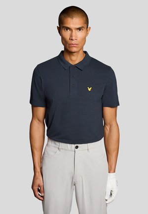 Mann trägt ein dunkelblaues Polohemd mit einem gelben Vogel-Logo, hellbeige Hose und einen weißen Handschuh an der rechten Hand vor einem einfarbigen Hintergrund.