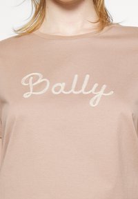 Personne portant une chemise beige avec le mot "Bally" brodé en lettres cursives blanches sur la poitrine.