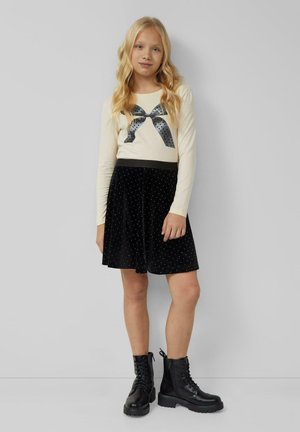 Crème lange mouw top met een zwarte stippenstrik graphic; zwarte fluwelen rok met witte stipdetails; gecombineerd met zwarte veterschoenen.