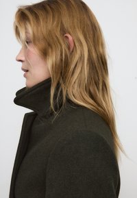 Cappotto scuro verde su misura con colletto alto, realizzato in tessuto testurizzato, che presenta un design aderente e linee pulite, mostrato in profilo.