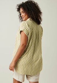 Blusa de manga corta, verde oliva con patrón geométrico blanco, tejido ligero, corte relajado, dobladillo curvado, sutil detalle de pliegue en la parte posterior.