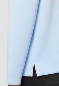 Lichtblauwe lange mouwen top met een licht gestructureerde afwerking. Heeft een gespleten zoom en zichtbare stiksels langs de manchet en naden.