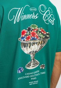 Zelené bavlněné tričko s stříbrným pohárem plným barevných pokerových žetonů a textem "Winners Club" nahoře, a detaily kasina dole.