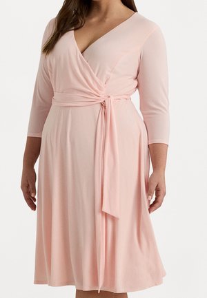 Frau trägt ein knielanges, hellrosa Wickelkleid mit Dreiviertelärmeln und gebundenem Gürtel in der Taille, stehend vor einem einfarbigen Hintergrund.