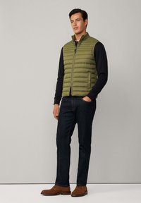 Olivgrüner, gepolsterter Weste mit hohem Kragen, Reißverschluss und seitlichen Taschen, kombiniert mit einem schwarzen Langarmoberteil und dunklen Jeans. Braune Schuhe.