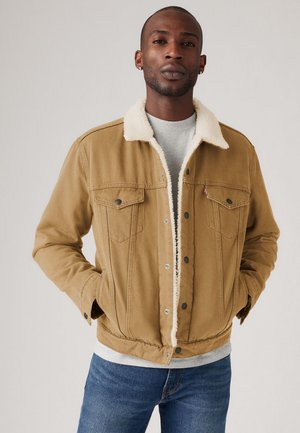 Tan denim jack met een witte fleece kraag, met twee borstzakken, drukknopen en een geribde tailleband. Draagt over een grijze shirt.