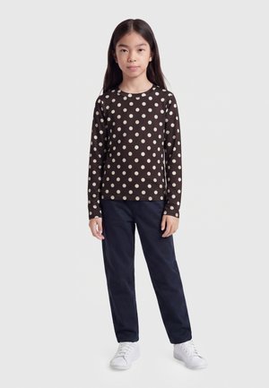 Fille debout portant une chemise marron à manches longues avec des pois blancs, un pantalon sombre et des baskets blanches sur un fond uni.