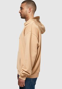 Beige hette laget av mykt stoff, med avslappet passform, kengurulommme, justerbar snorhette og ribbede mansjetter.