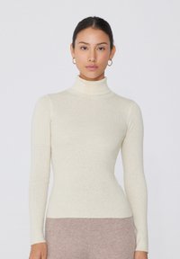 Hellbeige Rollkragenpullover mit geripptem Design und langen Ärmeln, figurbetont. Kombiniert mit beigen, hochgezogenen Hosen.
