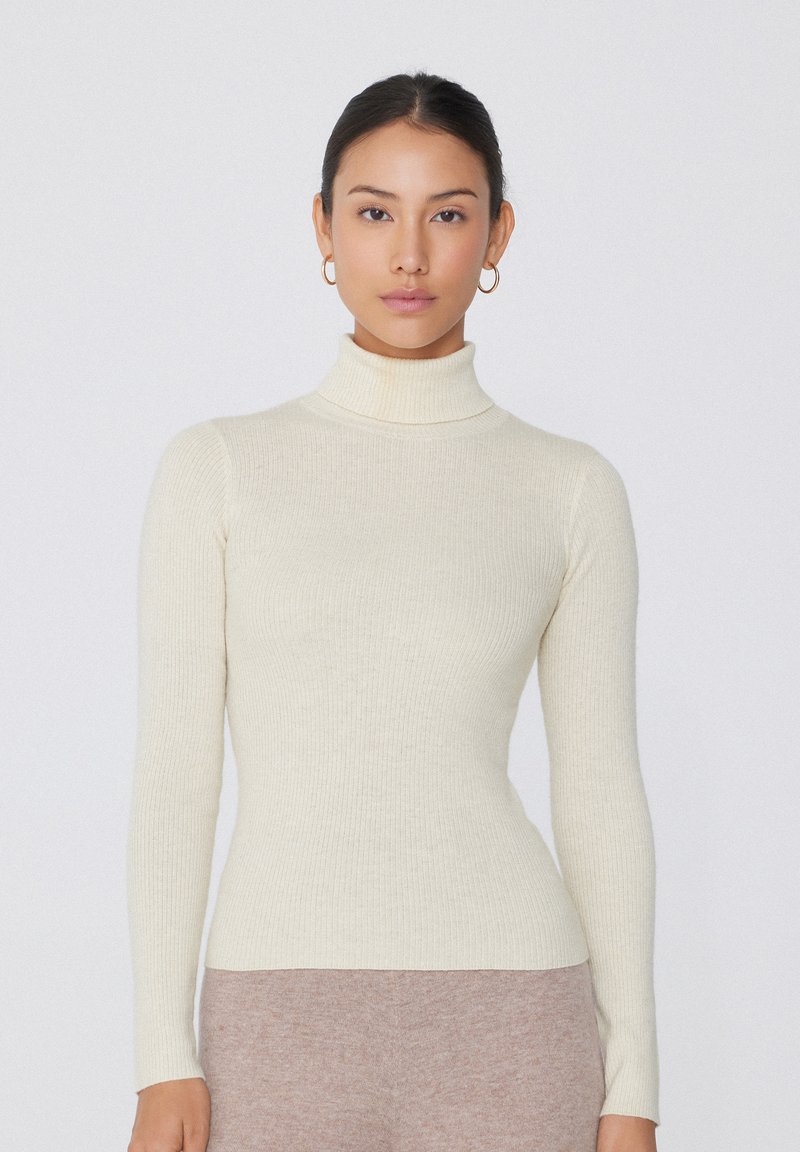Hellbeige Rollkragenpullover mit geripptem Design und langen Ärmeln, figurbetont. Kombiniert mit beigen, hochgezogenen Hosen.