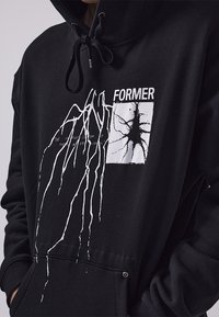 Svart hettegenser med frontlomme, med et hvitt grafisk trykk av abstrakte linjer og et firkantet design med tekst. Snorhette-detalj.