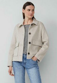 Beige wollen jas in een wolmix met een kraag, knoopsluiting en twee voorzakken, gedragen over een zwart-wit gestreepte shirt en blauwe spijkerbroek.