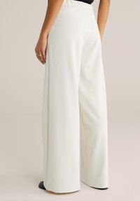 Sieviete valkā augsta vidukļa, plašu kāju baltas bikses, stāvot ar vienu roku atviegloti pie sāniem, apakšā redzamas melnas kurpes.