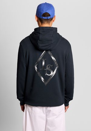 Person iført sort hættetrøje med reflekterende diamantformet "ls" logo på ryggen, blå kasket og hvide bukser, vendt væk mod ensfarvet baggrund.