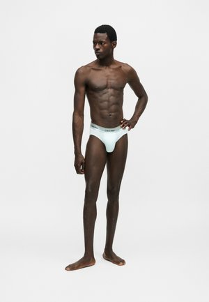 Male model with dark skin wearing light blue Calvin Klein briefs, standing barefoot with one hand on hip against white background.