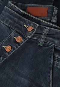 Dark denim jeans with a smooth texture, featuring copper-colored buttons and a brown leather tag labeled "TATUUM" on the waistband.