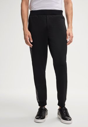 Pantalons de sport noirs avec une taille élastique, des poignets côtelés et des panneaux latéraux à motifs. Portés avec des baskets noires pour le contraste.