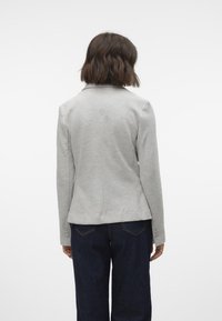 Šedý dobře padnoucí blazer s klopovým límcem, zadními švy a knoflíkovými detaily na rukávech, v kombinaci s tmavými džínami.