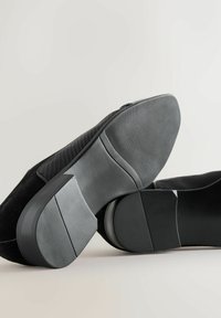 Schwarze Wildleder-Halbschuhe, schräg dargestellt, um die strukturierten Gummisohlen und niedrigen Absätze vor einem hellen neutralen Hintergrund zu zeigen.