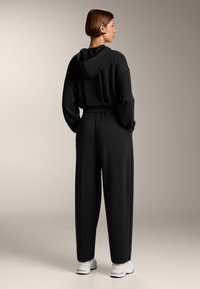 Felpa nera con vestibilità ampia, dotata di cappuccio e spalle basse. Abbinata a pantaloni neri a gamba larga e sneaker bianche. Tessuto liscio.