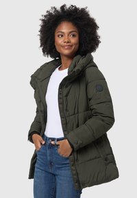 Doudoune vert olive avec capuche, design matelassé, boutons-pression et poches latérales. Associée à une chemise blanche et un jean bleu.