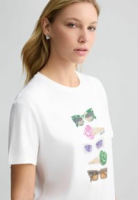 T-shirt en coton blanc avec des lunettes de soleil imprimées colorées et texturées ainsi qu'un design de main à l'avant, col rond et manches courtes.