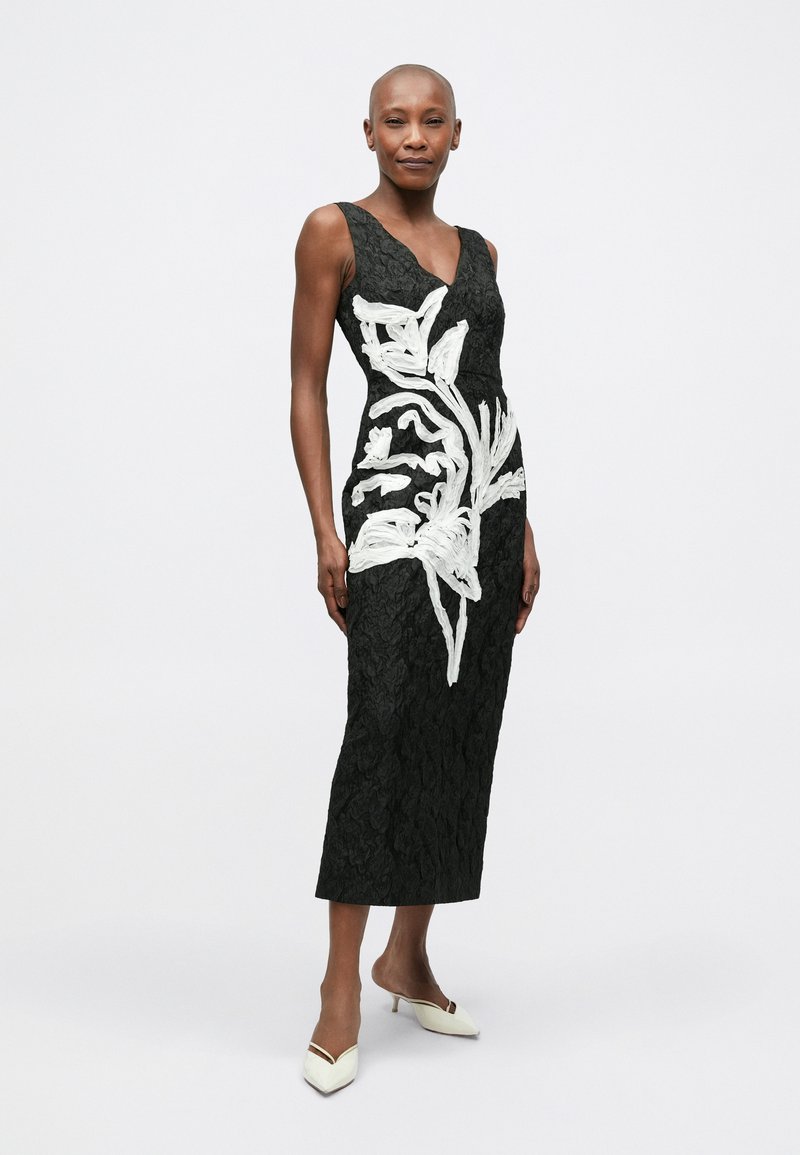 Black lace dress with white floral embroidery, deep V-neck, fitted waist, flared hem, and sleeveless design. Worn with white heeled shoes.