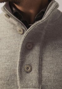 Grau gestrickter Pullover mit hohem Kragen, ausgestattet mit drei runden Knöpfen und einem strukturierten Strickmuster. Der innere Kragen zeigt einen karierten Stoff.
