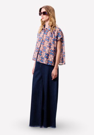 Vrouw met oversized vierkante zonnebril, een korte mouwen blauw-oranje geruit blouse en wijde donkerblauwe spijkerbroek.
