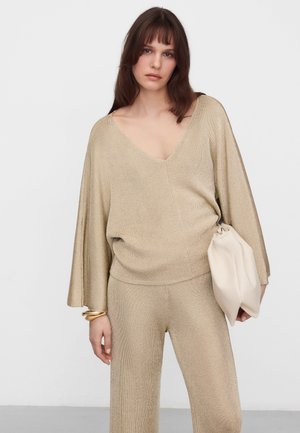 Donna che indossa un top beige lavorato a maglia larga con scollo a V e maniche larghe, abbinato a pantaloni coordinati, mentre tiene in una mano una morbida pochette beige.