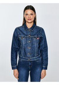 Sirgete tumedate juustega naine, kes kannab kuldseid rõngaskõrvarõngaid, nööbitud sinist teksajope ja sobivaid siniseid teksapükse, seistes ühevärvilise tausta ees.