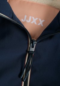 Chaqueta azul marino con la cremallera parcialmente abierta, mostrando el forro beige y una etiqueta de tela rosa con el logo blanco "JJXX" dentro del cuello.