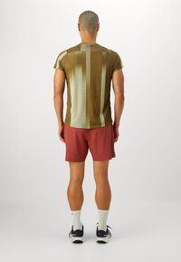 Mann som står med ryggen til, iført brun mønstrete kortermet skjorte, rustfargede shorts, hvite sokker og svarte joggesko på ensfarget bakgrunn.
