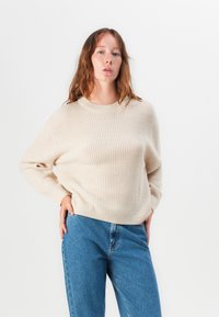 Pull en tricot beige avec un col rond et des manches longues, associé à un jean en denim bleu. La texture semble douce et côtelée.