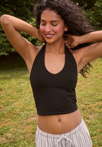 Crni halter top s dubokim dekolteom, napravljen od glatke tkanine. Nositi s laganim prugastim pidžama hlačama. Pozadina od zelene trave.