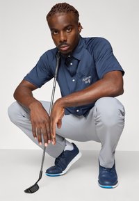 Marineblå polo med teksturert stoff og frontlomme, kombinert med lysegrå bukser og marineblå golf sko, mens han holder en golfklubbe.