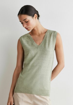 Kobieta ubrana w jasnozieloną bezrękawnikową kamizelkę z dekoltem w serek, stoi przy białej ścianie, patrząc w dół z rozluźnionymi ramionami.