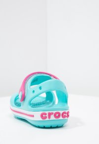 Lasten turkoosi ja vaaleanpunainen Crocs-sandaali, jossa on kantahihna ja teksturoitu pohja, aseteltuna valkoiselle pinnalle yksinkertaista taustaa vasten.