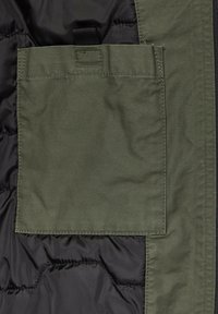 Grünes Stofftasche auf einer schwarzen, gesteppten Jacke; mit einem Klappenverschluss und sichtbarem Nähen, das eine kontrastierende Textur bietet.