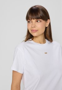 Biała koszulka z krótkim rękawem z bawełny, z małym złotym akcentem na piersi. Gładka tekstura z klasycznym okrągłym dekoltem.