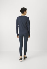 Sötétkék, hosszú ujjú sportfelső ívelt aljjal és raglán ujjal, sötét leggingsel párosítva. A lábbeli fekete-fehér dizájnnal rendelkezik.
