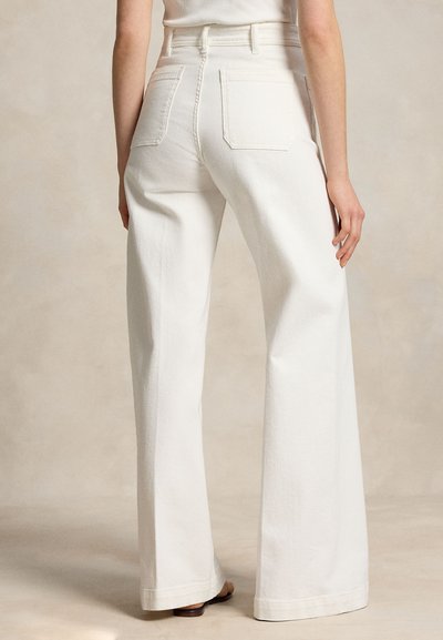 Γυναίκα που φορά λευκά τζιν με ψηλή μέση, φαρδιά πόδια και πίσω τσέπες, όρθια, ξυπόλητη σε φωτεινό ουδέτερο φόντο.