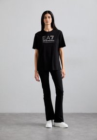 Melns kokvilnas t-krekls ar izteiksmīgu sudraba "EA7 EMPORIO ARMANI" logotipu, apvienots ar melniem flared biksēm un baltiem sneakeriem. Gluda tekstūra.