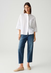 Witte oversized, knoopsluiting shirt met een hoge hals en een borstzak, gecombineerd met donkerblauwe losse jeans en beige flats.