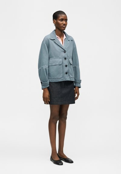 Femme debout portant une veste bleue boutonnée, une chemise à col rose, une jupe foncée et des ballerines noires, sur un fond clair uni.